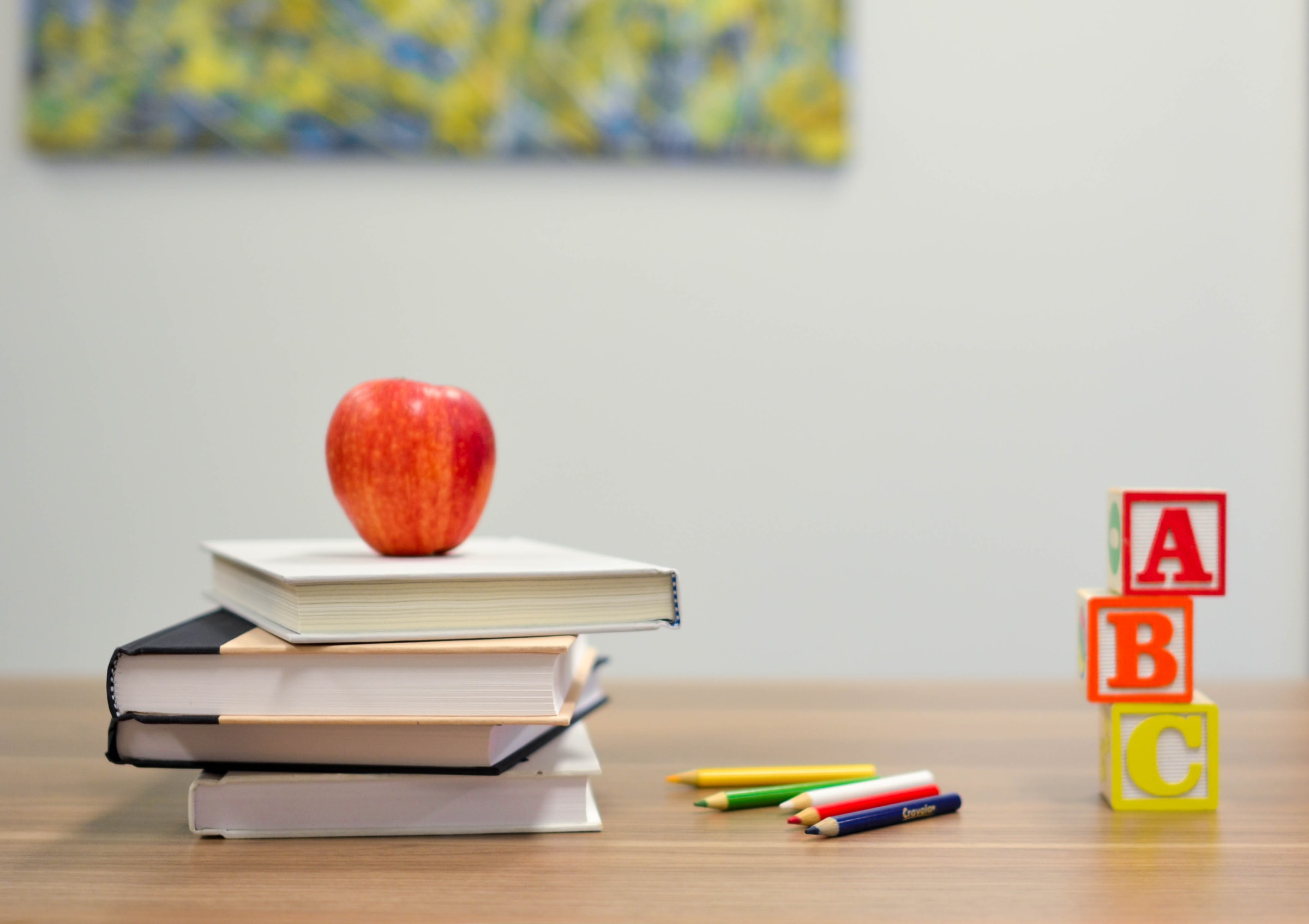 Image contains a stack of books with an apple on top, a row of colored pencils, and letters A, B, and C blocks vertically stacked.