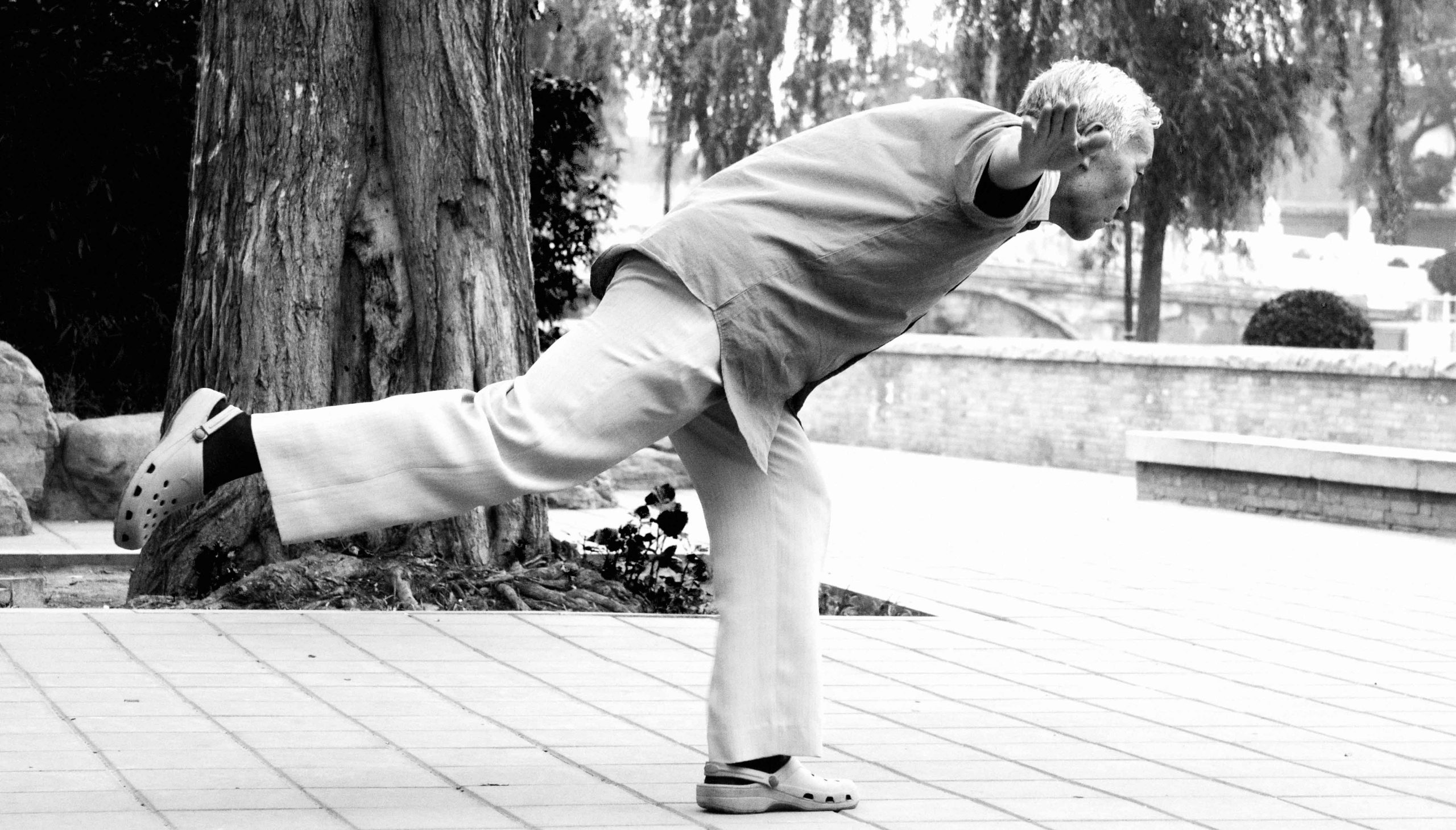 Older man balancing on one foot outdoors.