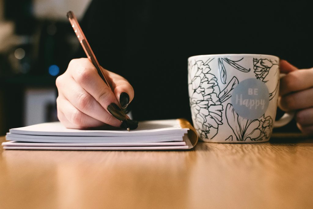 decorative photo shows a hand holding a pen writing in a journal while the other hand holds a mug saying "Be happy"