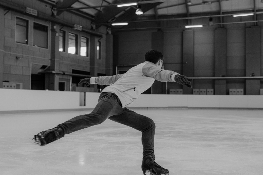side angle photograph of a male figure skater with arms out to the side, one leg back, and one skate on the ground