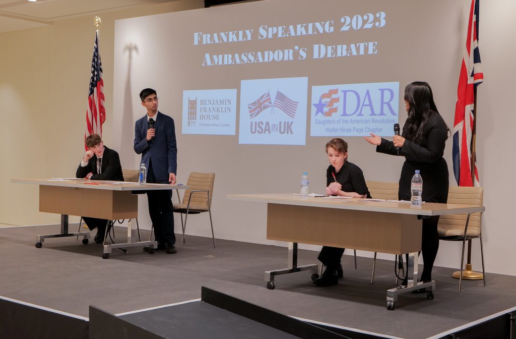 a photo showing four students behind two desks on a stage, two of the students standing and facing each other holding microphones, with the words "Frankly Speaking 2023 Ambassador's Debate" on the screen behind them