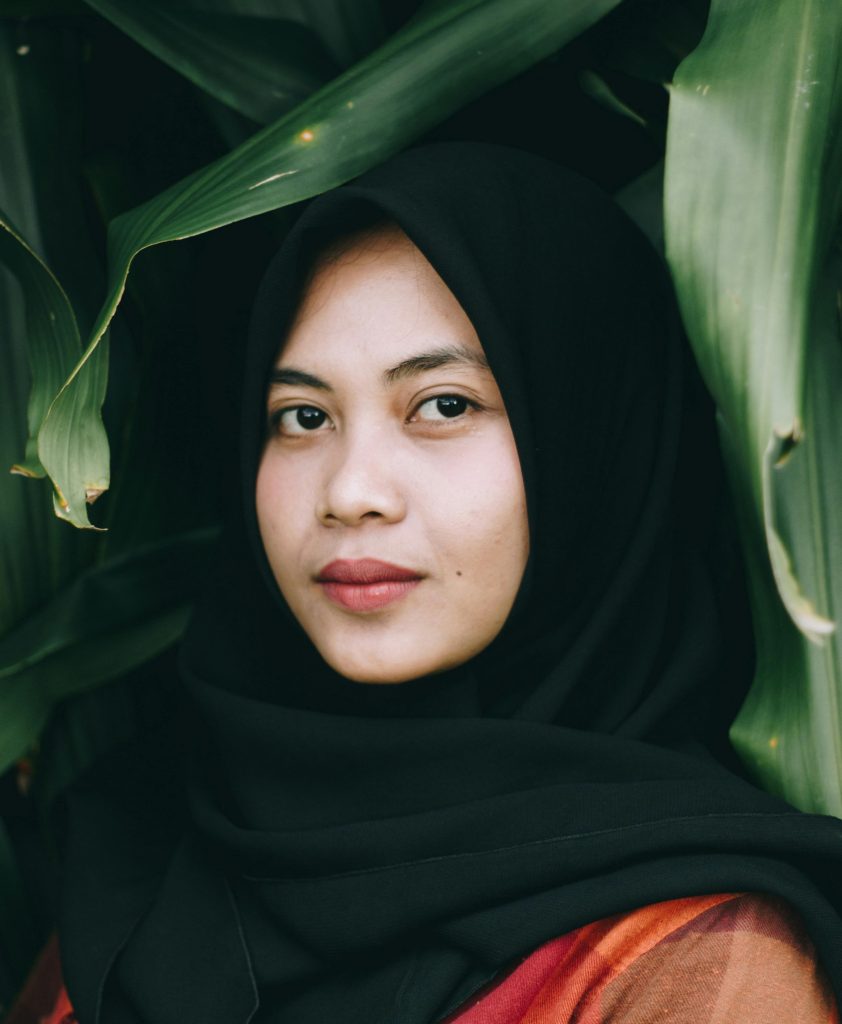 a photo of a woman with a black scarf wrapped over her head and around her neck to conceal her hair stands in front of large leaves