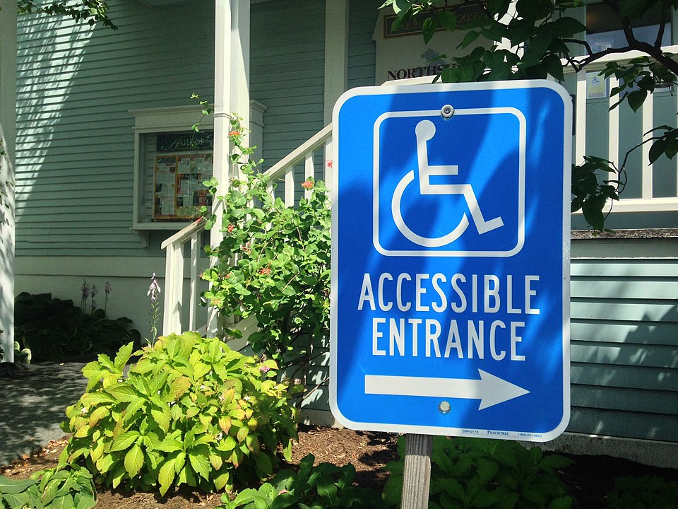 a photo of a blue sign with a wheelchair image and the words "accessible entrance" points to the right while the background of the image shows a set of stairs into an old building