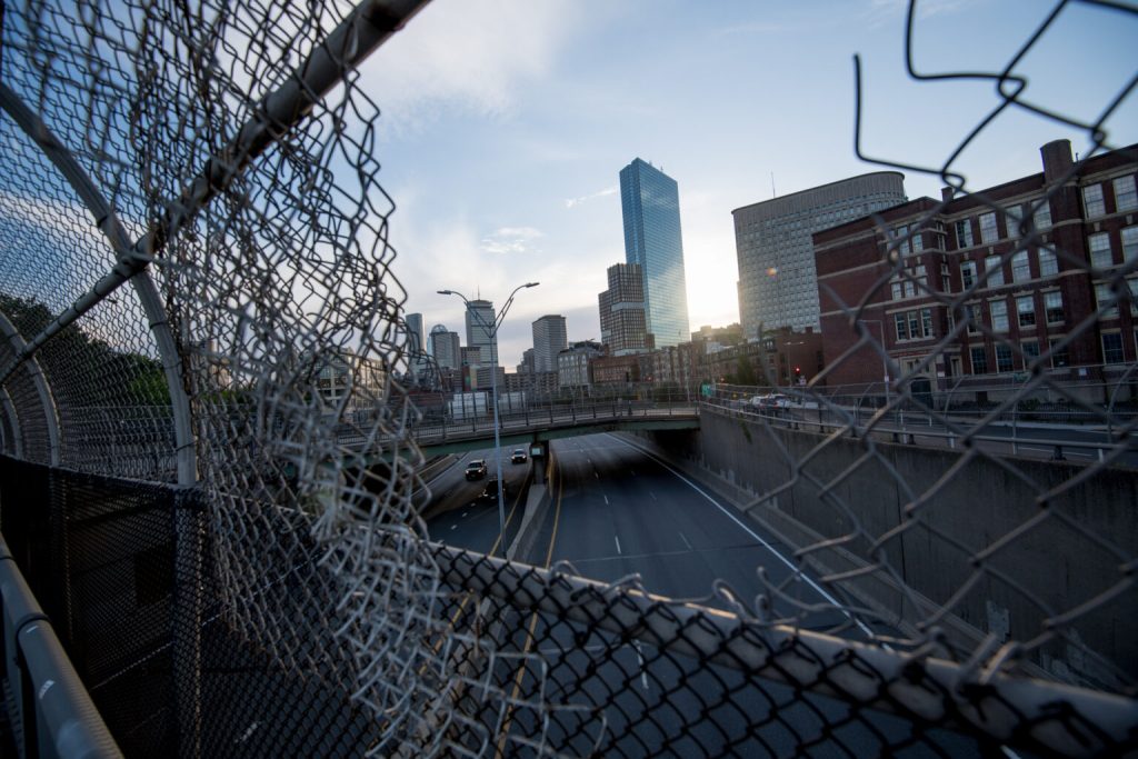 foreground of photo shows a large gaping hole in a chain link fence because many of the links are broken, revealing streets and city buildings in the background
