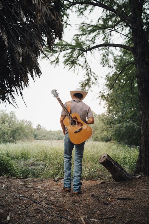 photograph of a man wearing a cowboy hat with a guitar on his back walks away from the camera into a forest meadow