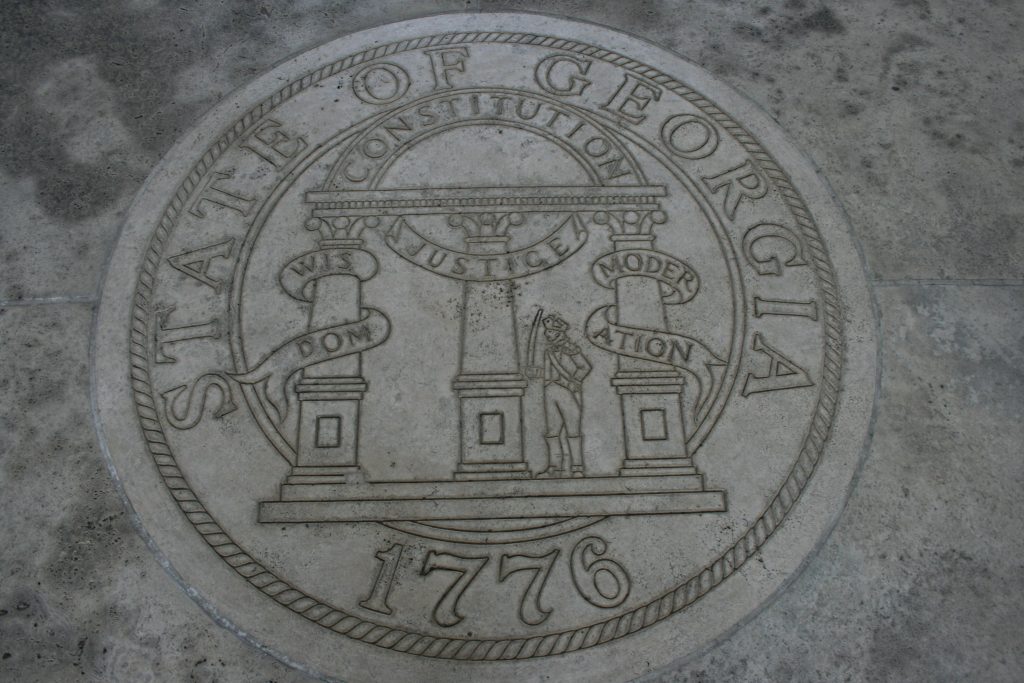 A picture of the Seal of the state of Georgia (dated 1776), engraved into stone. The description on the seal has three columns labeled “wisdom,” “justice,” and “moderation,” with a semi-circle labeled “constitution connecting the three.