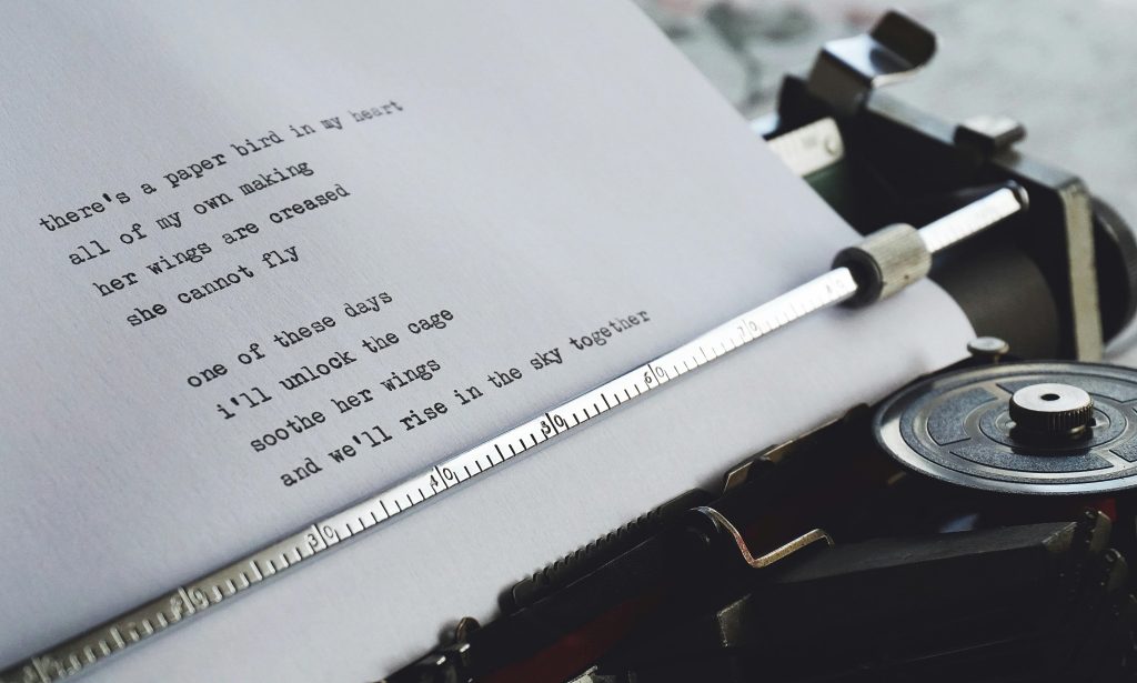 photo of a typewriter with a poem on the page that reads "there's a paper bird in my heart / all of my own making / her wings are creased / she cannot fly / one of these days / i'll unlock the cage / soothe her wings / and we'll rise in the sky together"