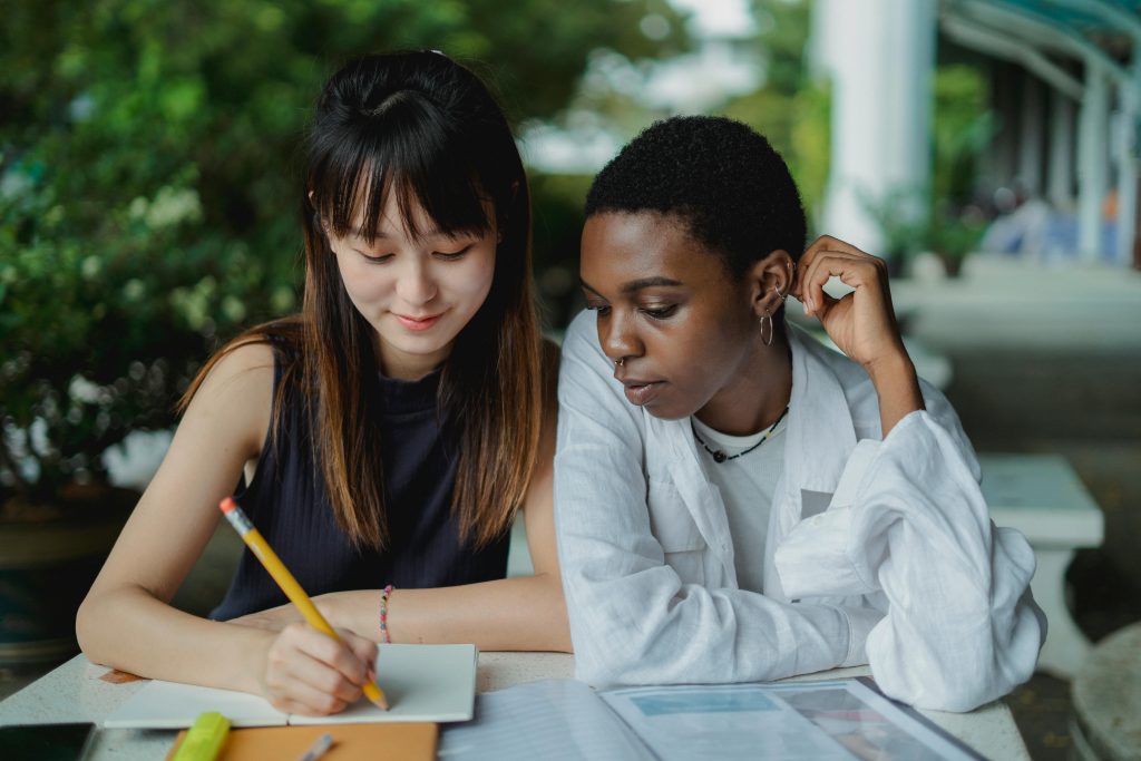 a photo of two college students, one writing while the other watches, interested