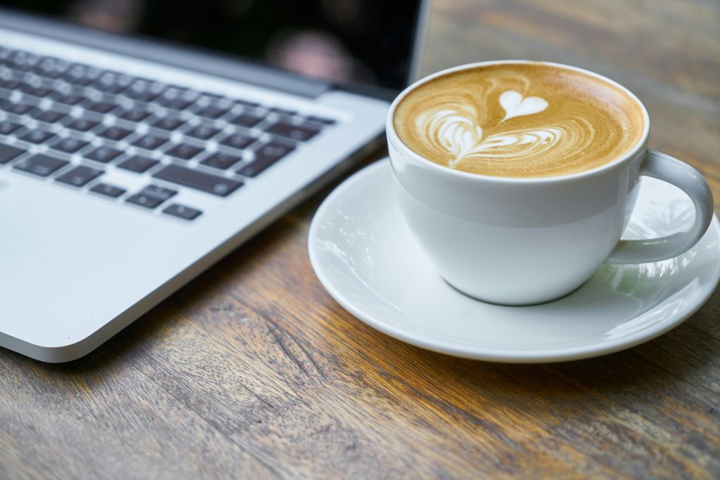 photo of a coffee mug with a heart swirled into the foam and set next to a laptop computer