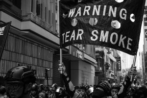 Black and white photo of protestors marching through the streets equipped with eye and head protective gear. One person is holding a banner that warns of tear smoke.