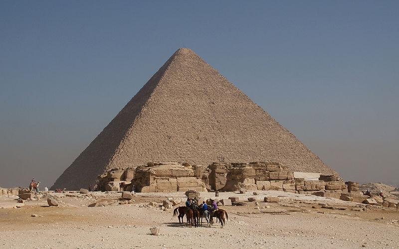Image of the Great pyramid shows people with horses in the foreground, a person riding a camel to the left side of the pyramid, and the structure itself rising from the desert floor towards a hazy sky in the background.