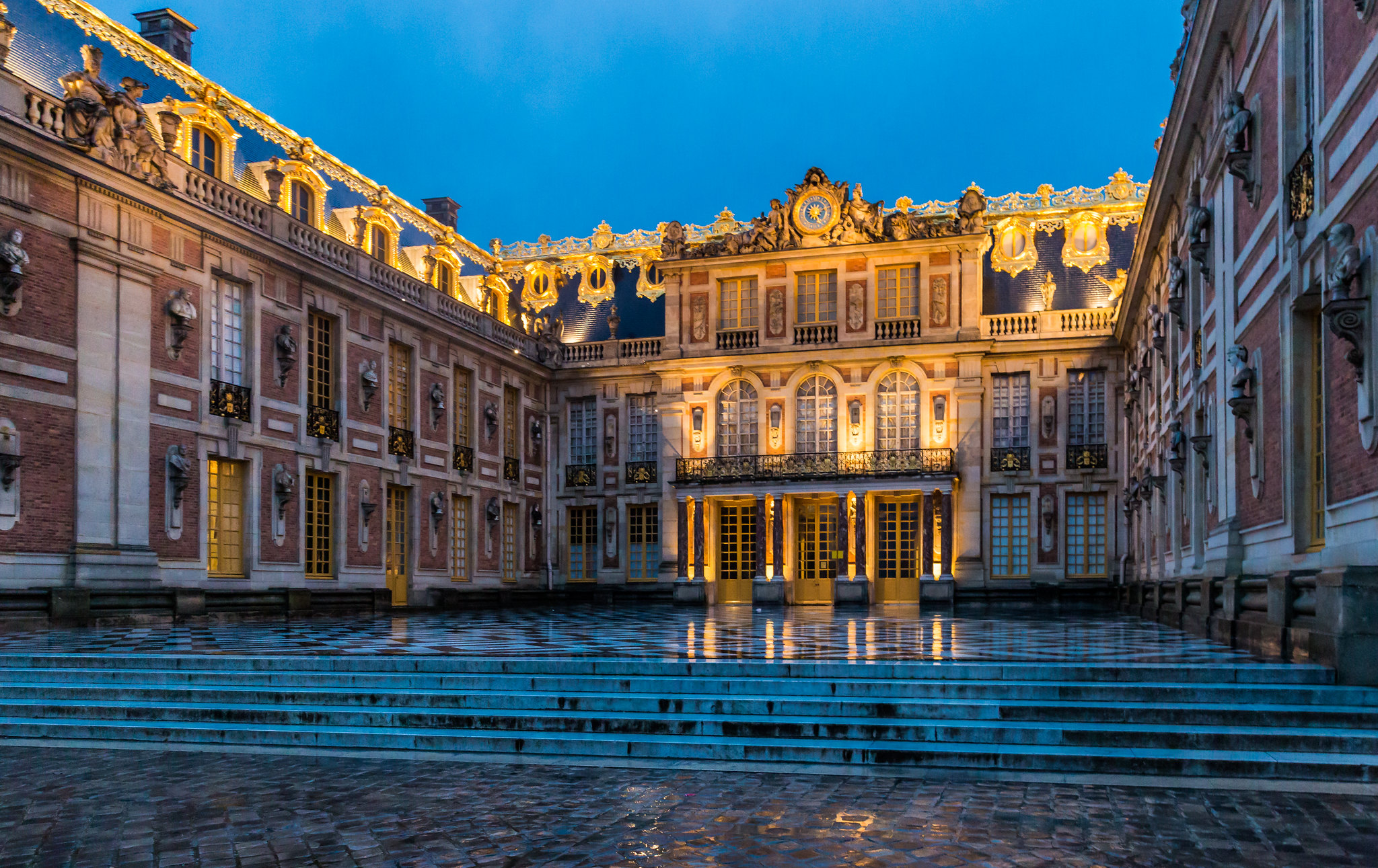 Vew of a courtyard paved with marble. The palace is lit as though it is dawn or dusk and the ornate walls and windows glow with light.